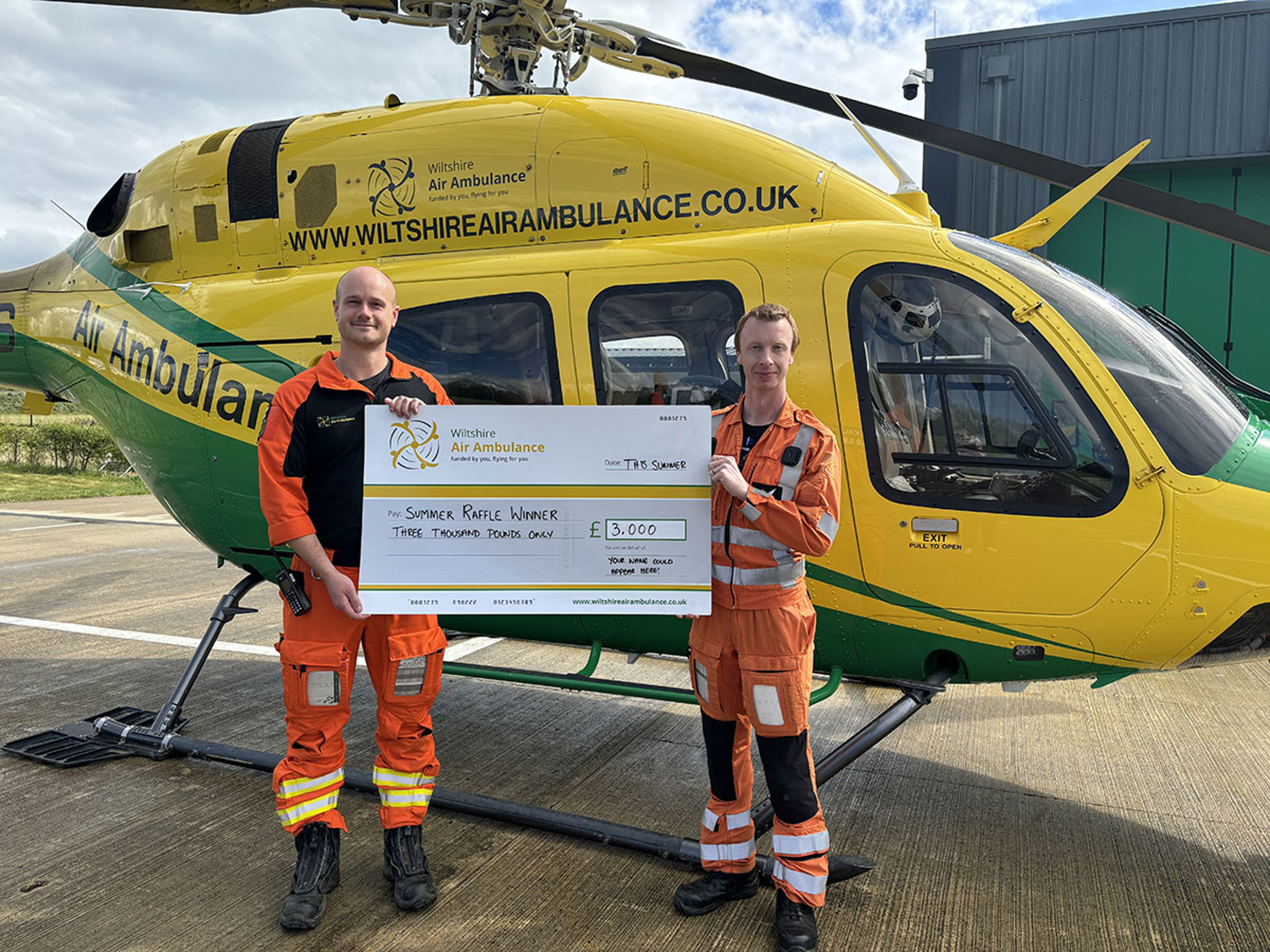 Two male paramedics, wearing orange flight suits, standing in front of a yellow and green helicopter, holding a large novelty cheque