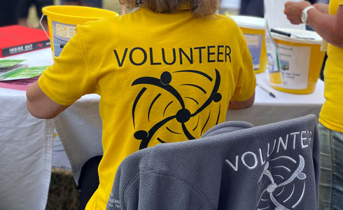 A volunteer at an event wearing a yellow t-shirt, they are sat at a table with yellow collection buckets on and raffle tickets