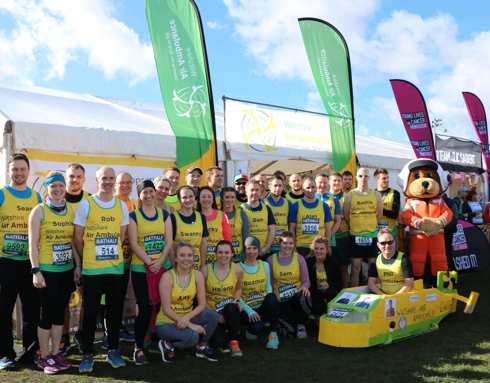 A group photo of participants together before running the Bath Half Marathon. All runners are wearing yellow and green running vests and are stood next to a large white gazebo.