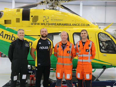 A group photo of two paramedics wearing orange flight suits and a pilot wearing a black flight suit with new ambassador Tom Dunn.