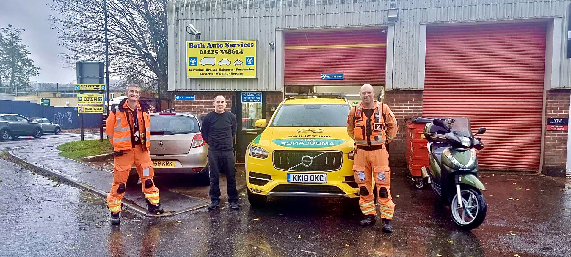 Critical care doctor Reuben and paramedic Dan wearing orange flight suits, standing in front of Bath Autos with a mechanic and Wiltshire Air Ambulance's critical care car