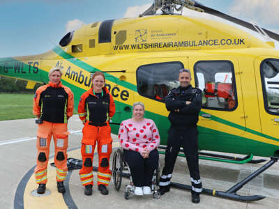 Two paramedics wearing orange flight suits and a pilot wearing a black flight suit stood with new ambassador Louise Hunt in front of the helicopter on a sunny day.