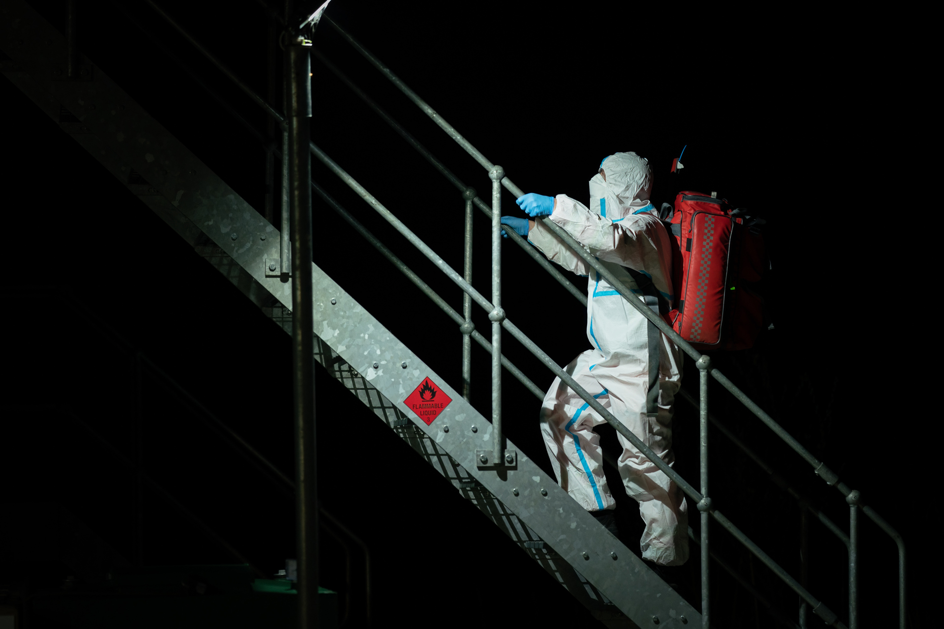 A photo of a paramedic in full hazmat PPE ascending metal stairs with a red kit bag on their back.