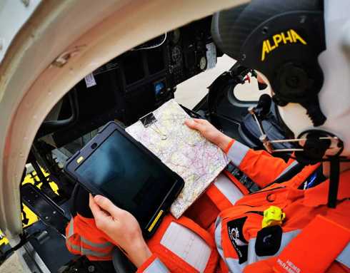 A paramedic sat in the cockpit of the helicopter looking down at a black iPad and a paper map. They are wearing a white and black flight helmet and orange flight suit.