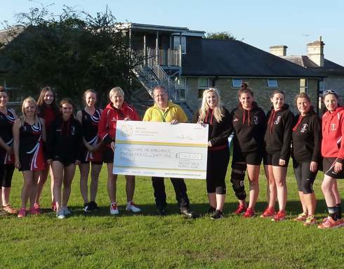A large group photo of netball players alongside a volunteer wearing a yellow polo shirt. They are collectively holding a giant novelty cheque.