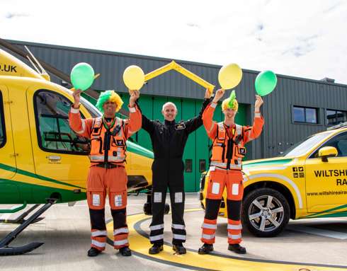 Three members of Wiltshire Air Ambulance aircrew wearing yellow and green wigs and holding balloons in front of the Bell-429 helicopter and critical care car.