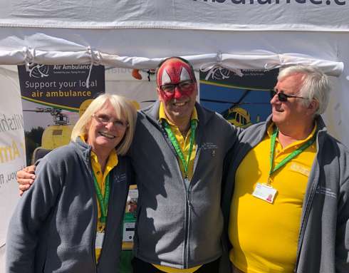 A group of three Wiltshire Air Ambulance volunteers wearing yellow polo shirts outside a gazebo at a fundraising event.