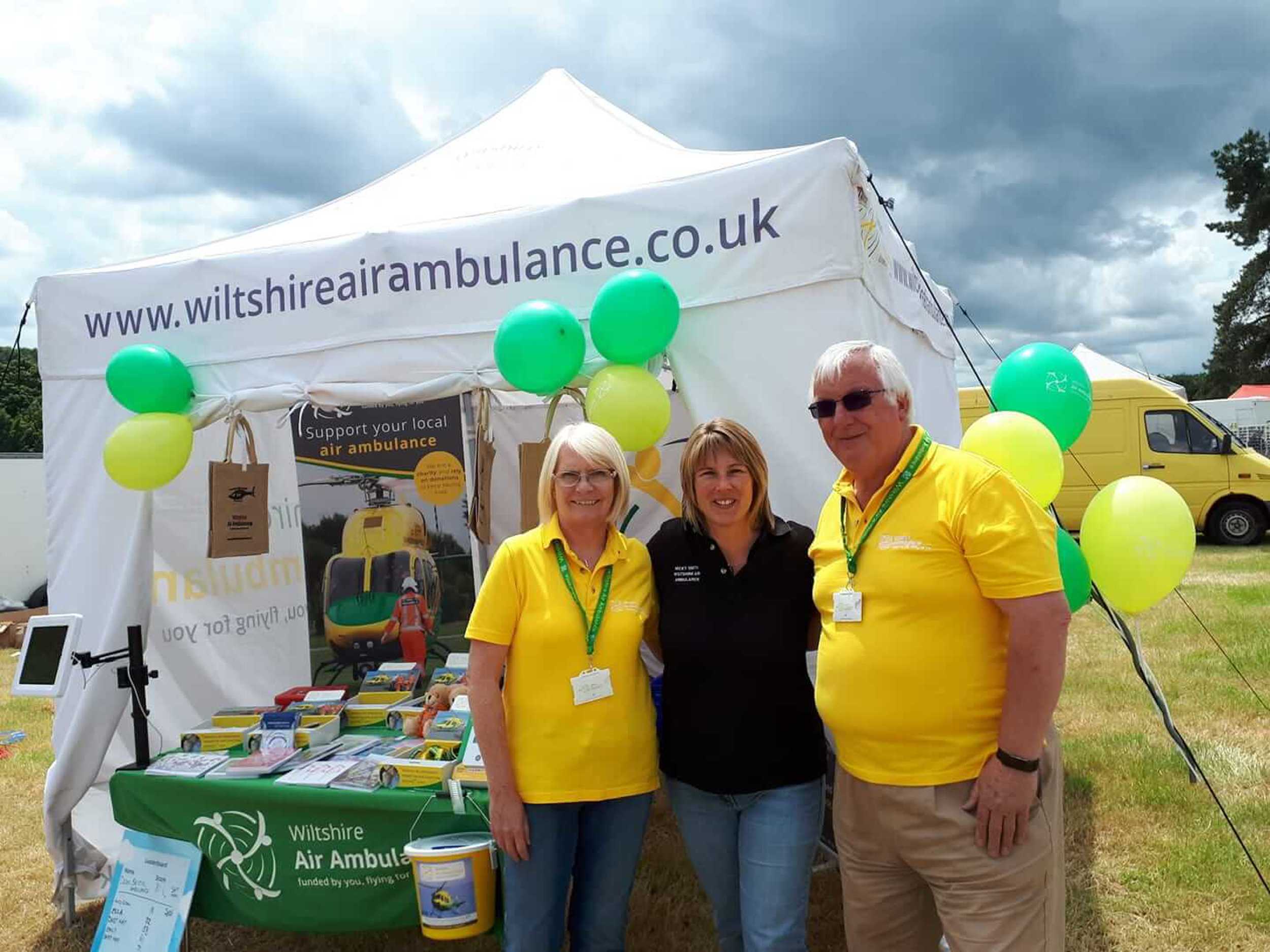 A photo of two Wiltshire Air Ambulance volunteers at an event with a Wiltshire Air Ambulance pilot. The photo is in front of a white gazebo with event merchandise.