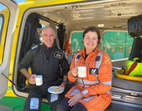 A pilot and paramedic smiling posing with a plate of tarts and holding mugs in the Wiltshire Air Ambulance helicopter