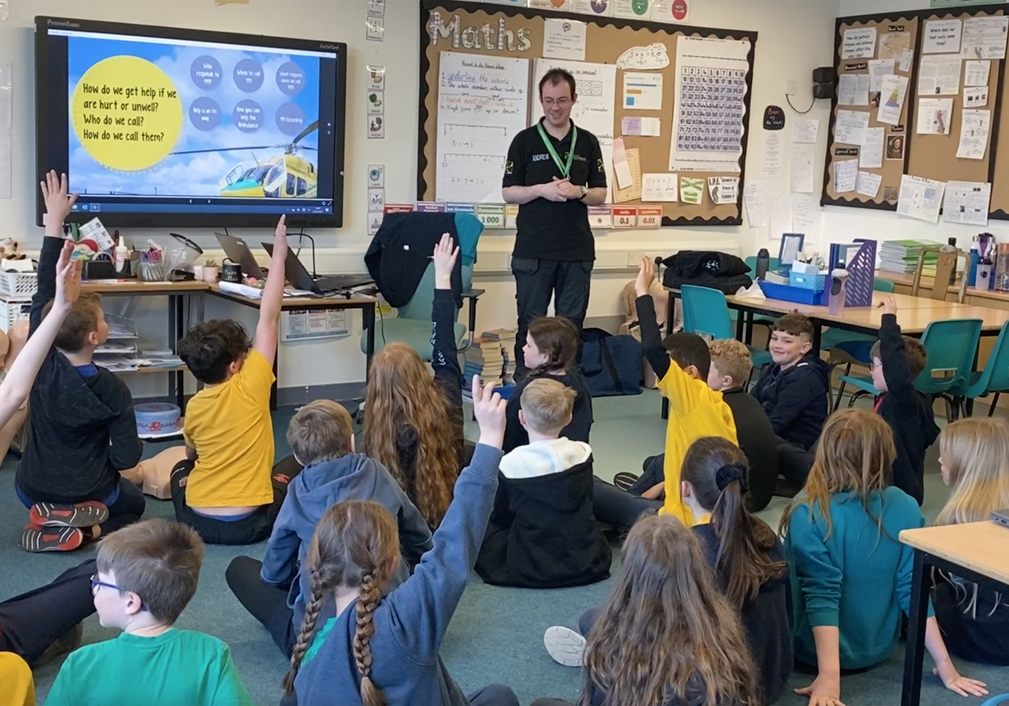 Trainer delivering emergency awareness training to a class of school children, who are sat on the floor with their hands up