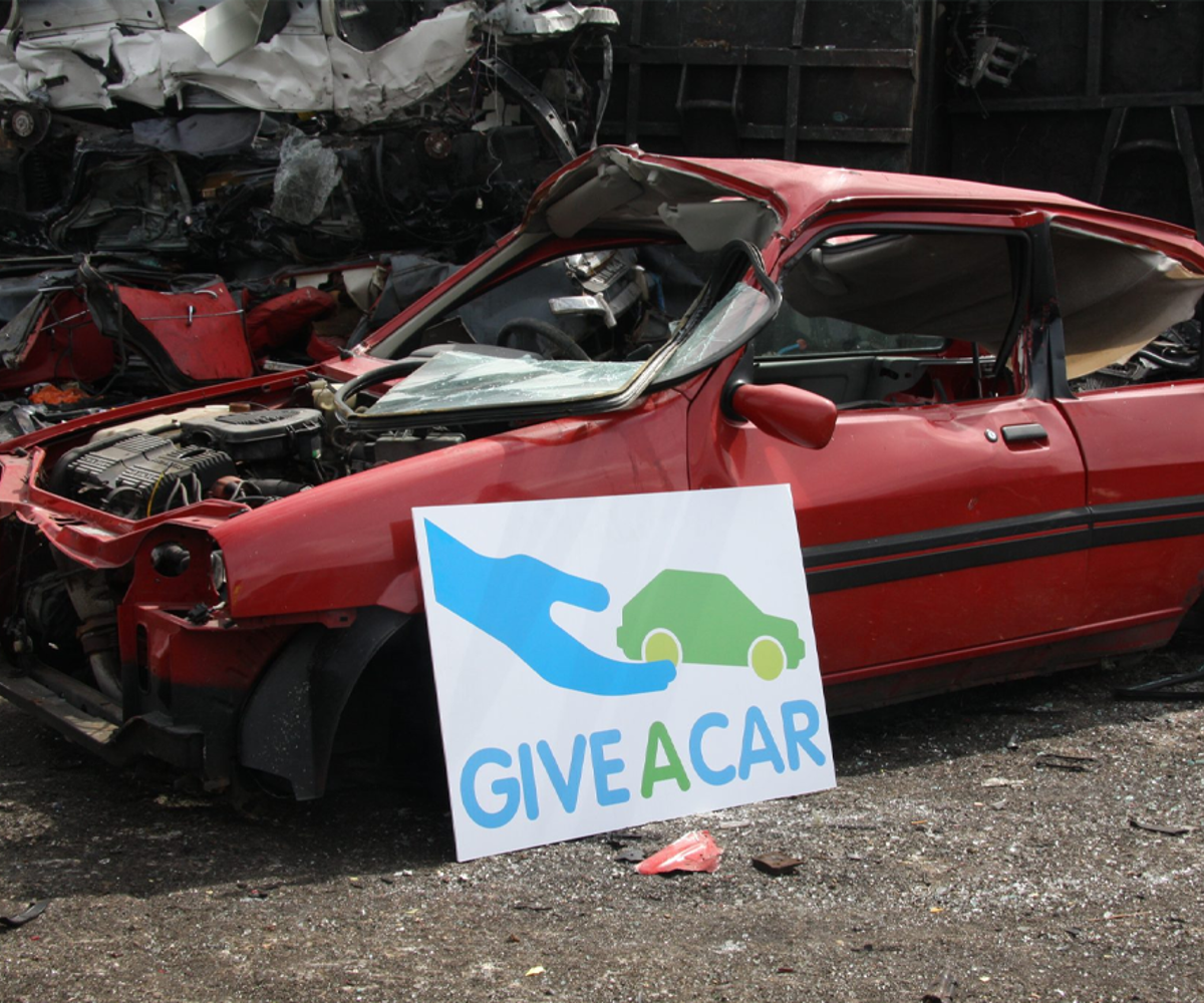 A scrapped red car behind a white board with the logo for Give A Car