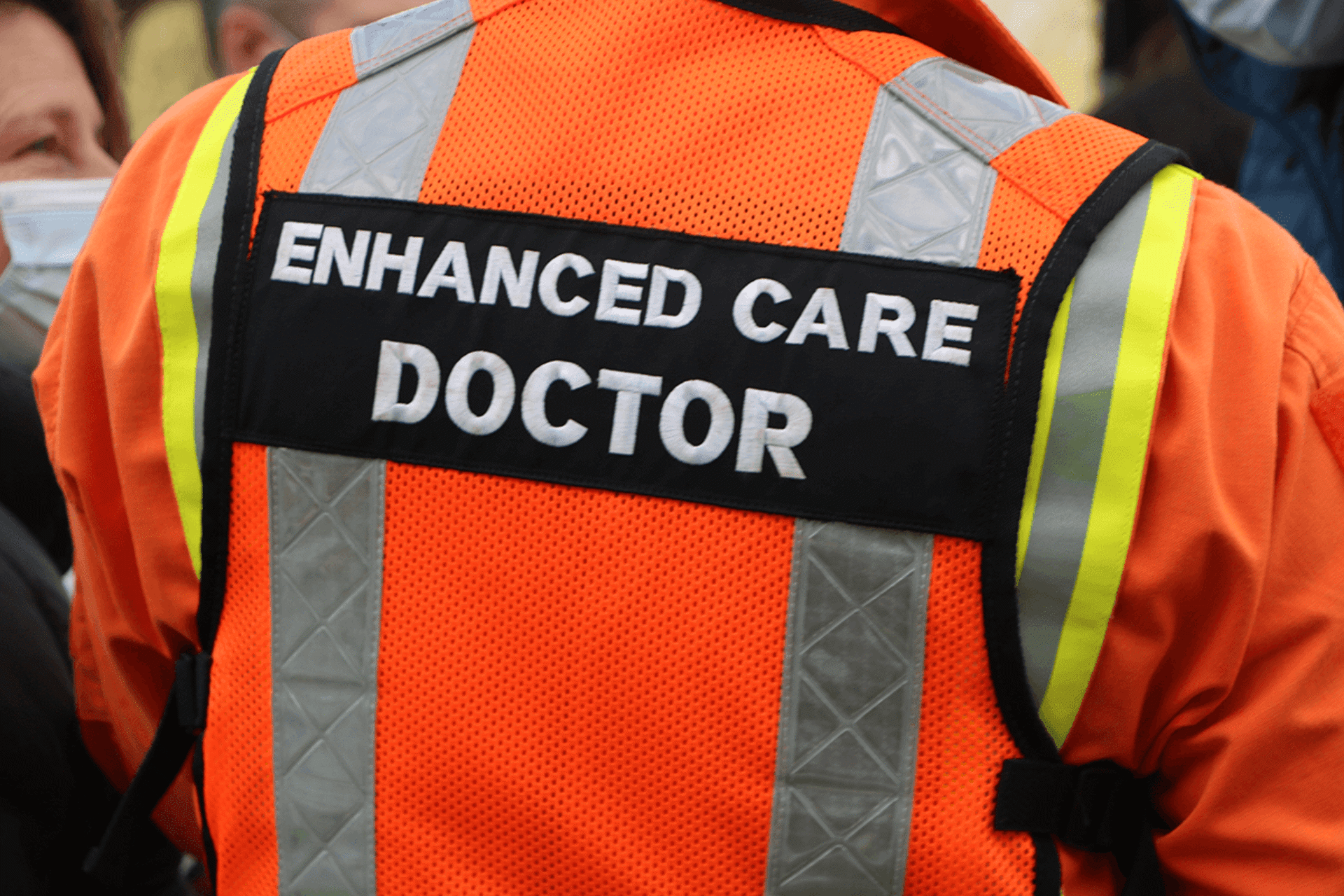 A close up of a person's back wearing and orange flight suit and vest with 'enhanced care doctor' written on the back