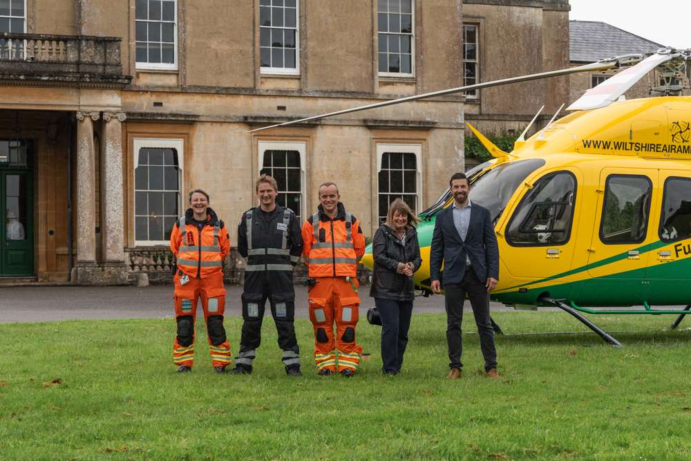 Two crew wearing orange, a pilot and two adults standing in front of a yellow and green helicopter in front of Hartham Park house