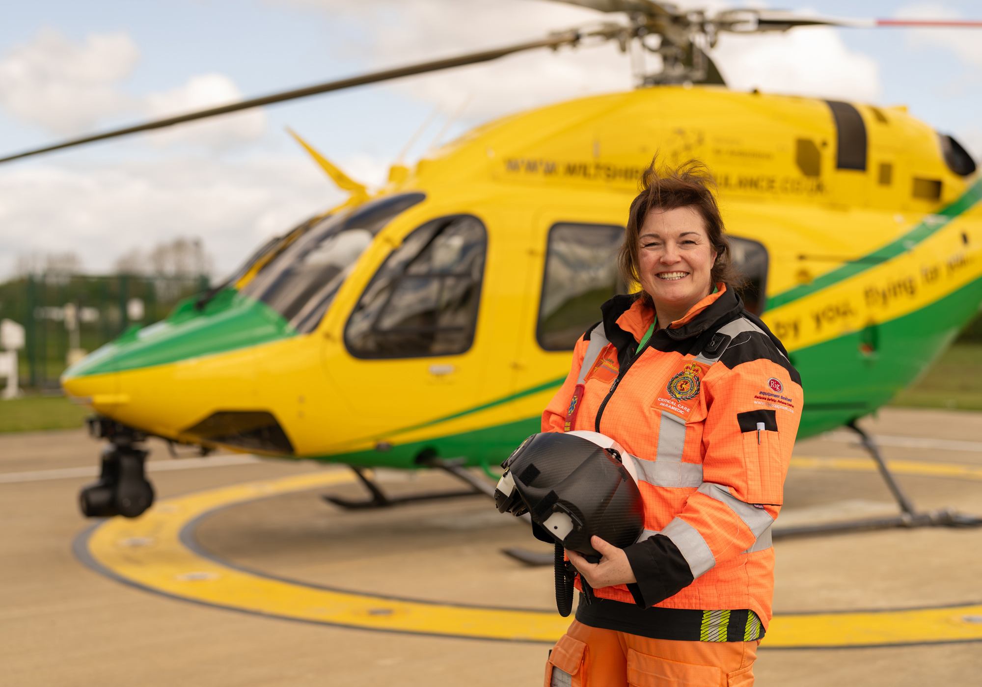 A photo of a female critical care paramedic holding a helmet, standing in front of a yellow and green helicopter