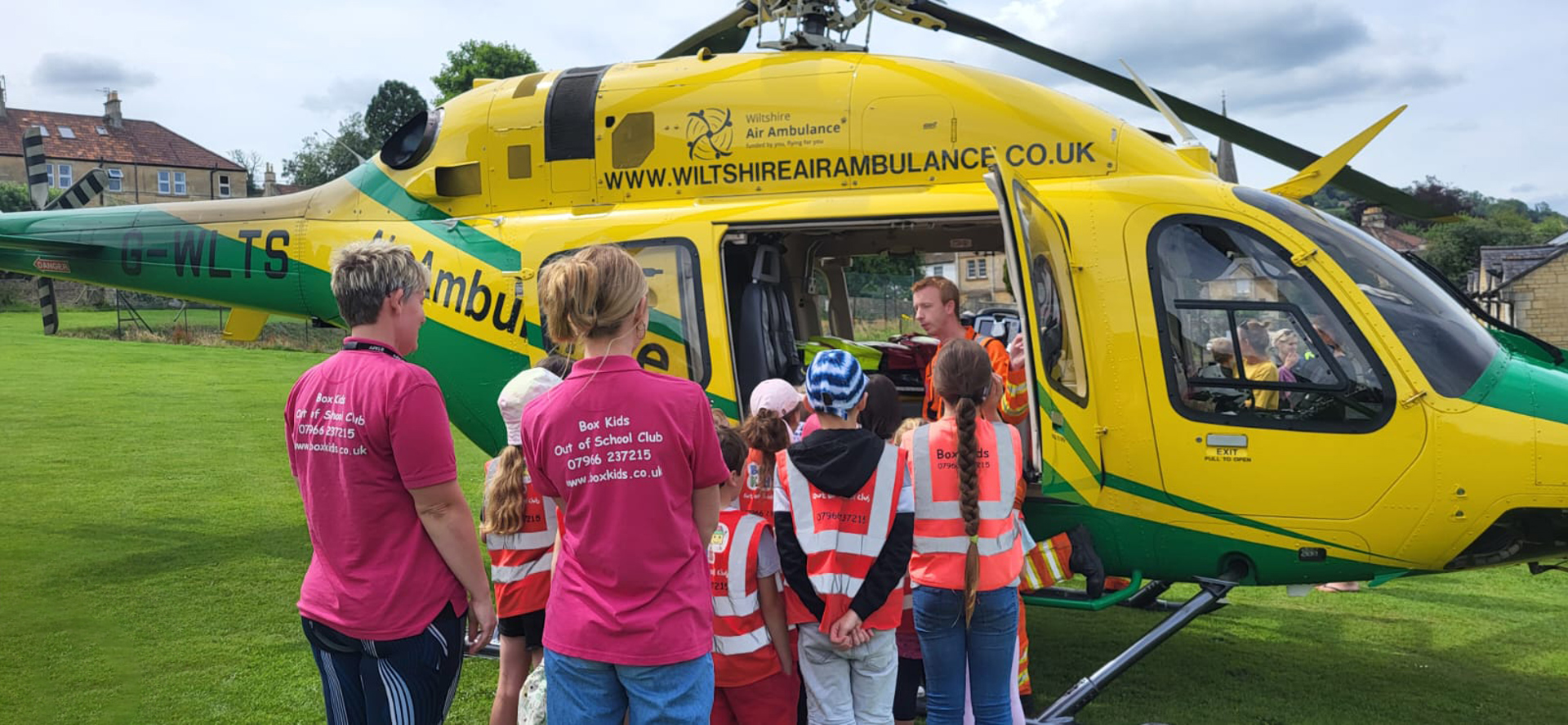 Children wearing high vis, accompanied by two adults, surrounding a yellow and green helicopter